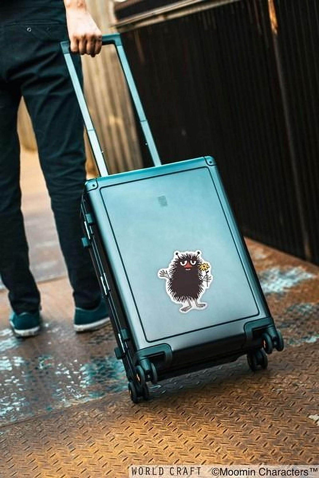 A person is pulling a suitcase with a sticker of a character on it, standing on a metal platform.