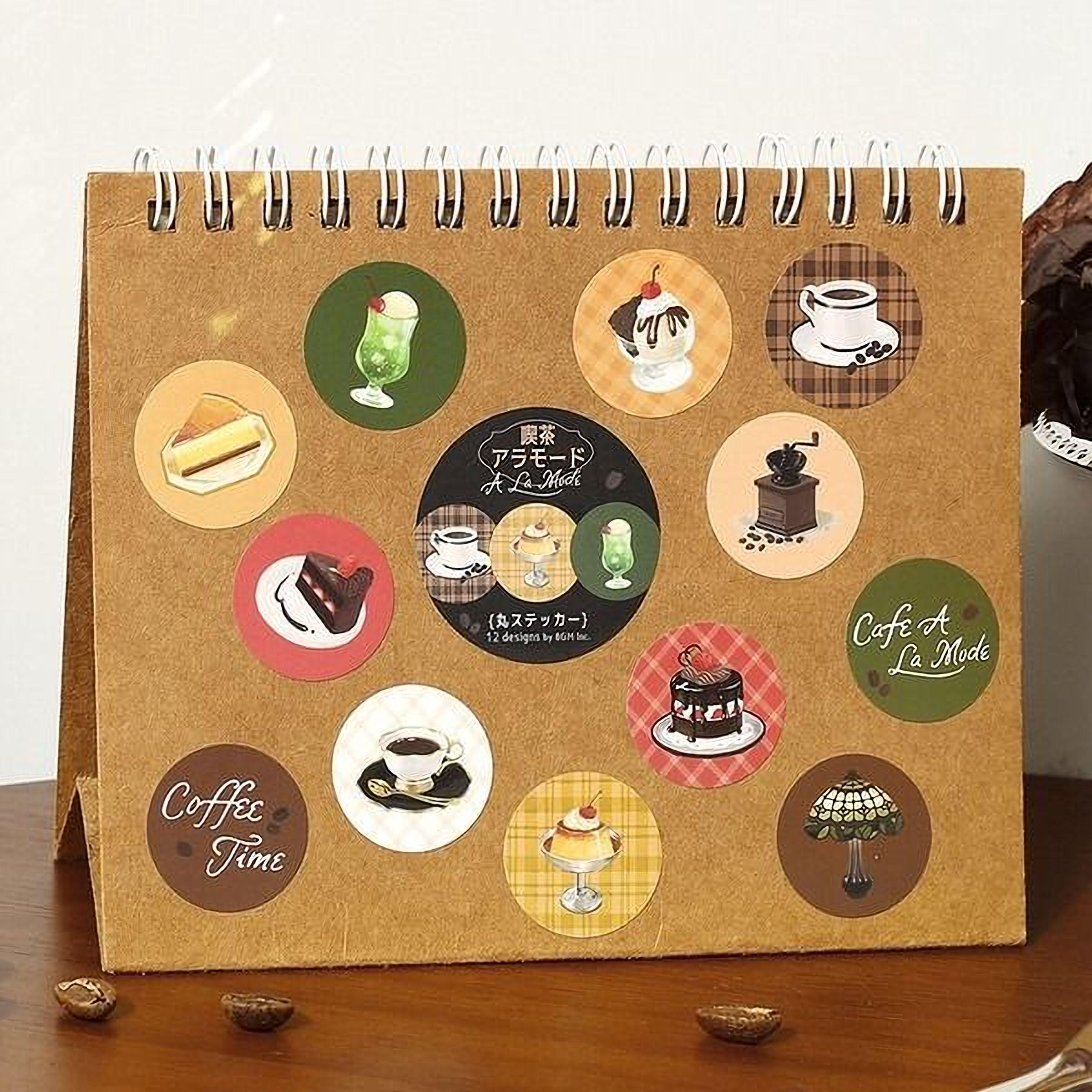 A spiral-bound notebook with various circular stickers depicting different food and drink items, such as a slice of cake, a cup of coffee, and a coffee machine.