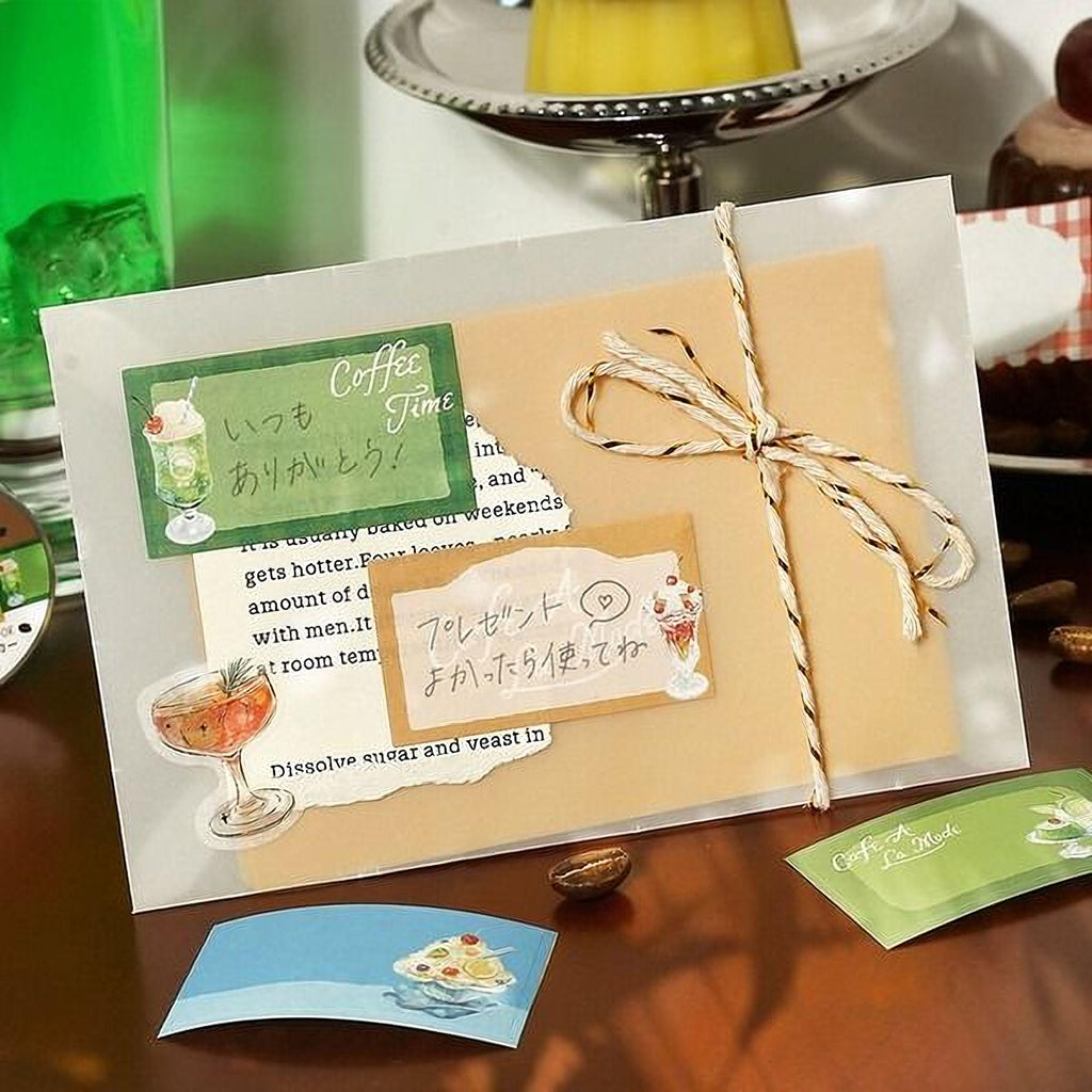 a card with various decorative elements, including a green card with a picture of a glass of wine, a yellow card with a picture of a cup of coffee, and a brown envelope with a picture of a cup of coffee. The card is placed on a wooden surface, and there are some coffee cups and other items in the background.