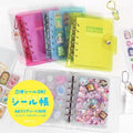 A collection of colorful notebooks and stickers, including a yellow sticker book and a pink notebook, along with various stickers and charms.