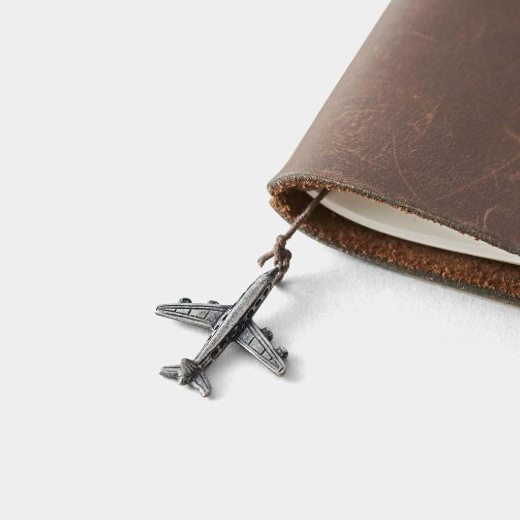 A small silver airplane charm is attached to a leather-bound notebook.