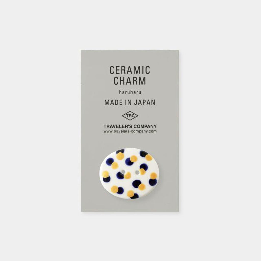 The image is a product advertisement for ceramic charms, specifically the "Ceramic Charm Haruharu Made in Japan". The product is described as a "Ceramic Charm" and is made in Japan. The company is Traveler's Company, and the website is www.travelers.com. The product is a round, white ceramic charm with blue and yellow dots.