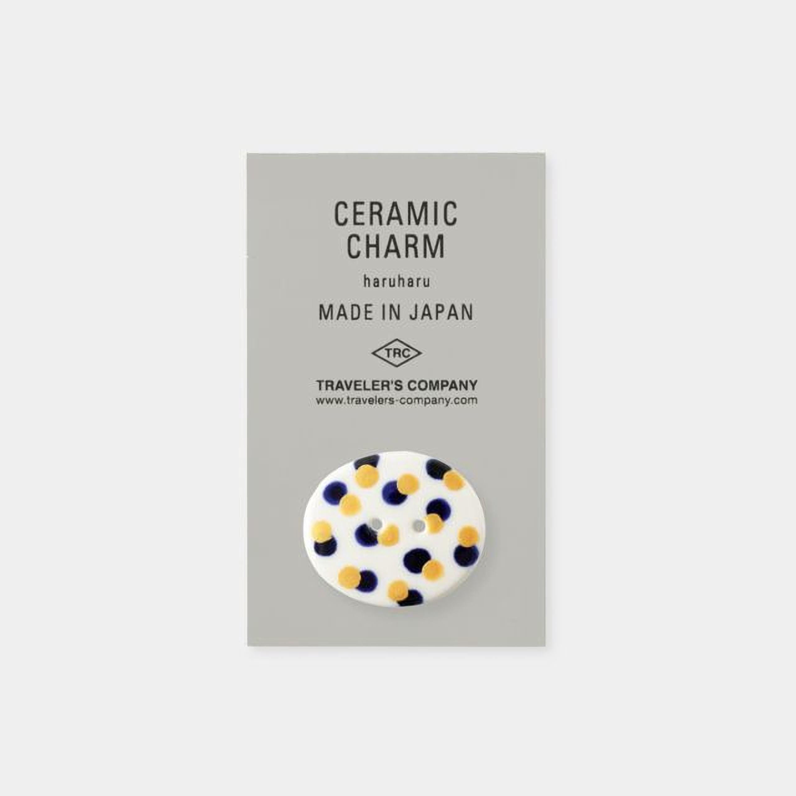 The image is a product advertisement for ceramic charms, specifically the "Ceramic Charm Haruharu Made in Japan". The product is described as a "Ceramic Charm" and is made in Japan. The company is Traveler's Company, and the website is www.travelers.com. The product is a round, white ceramic charm with blue and yellow dots.