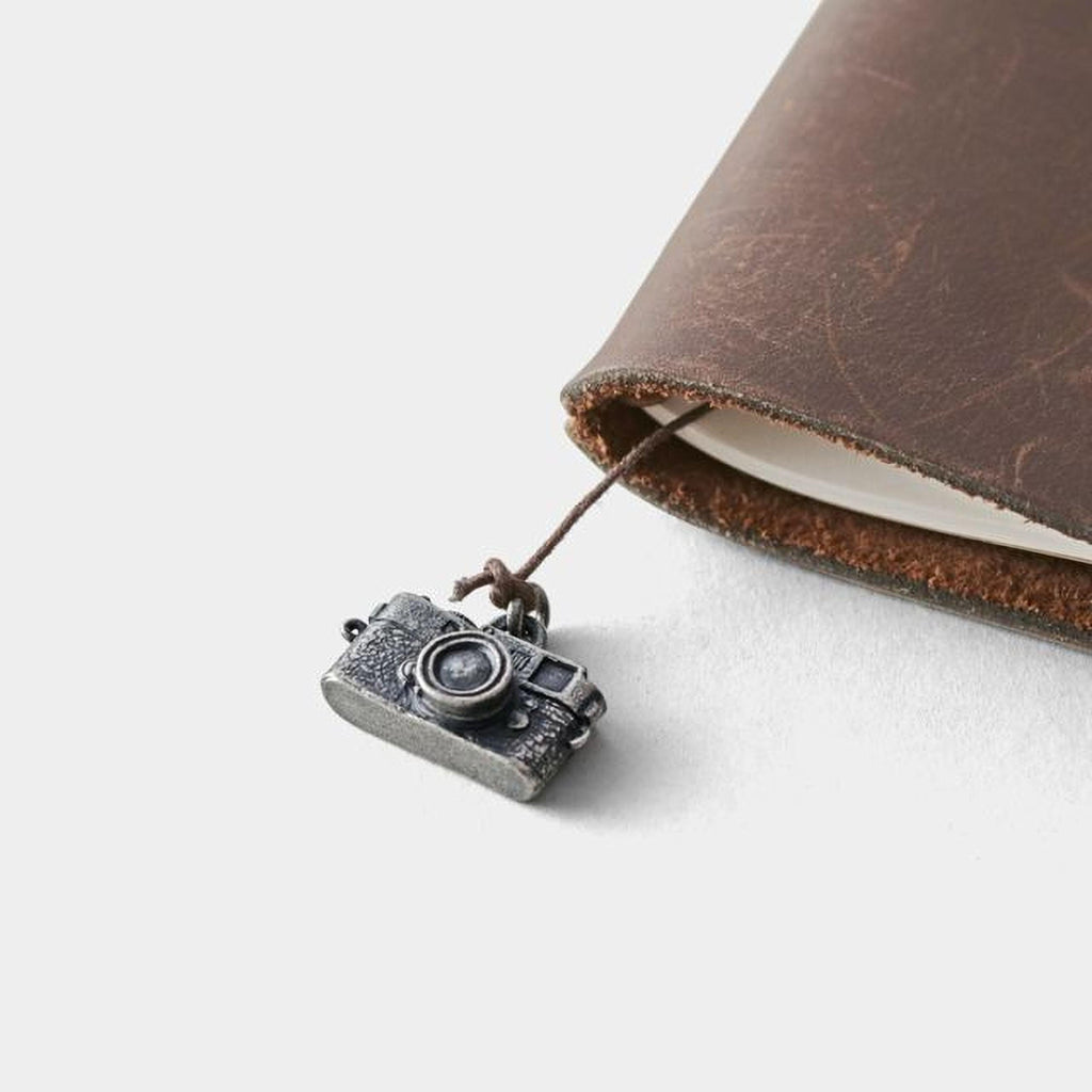 A small metal camera charm is attached to a leather-bound notebook, creating a visually appealing and organized composition.