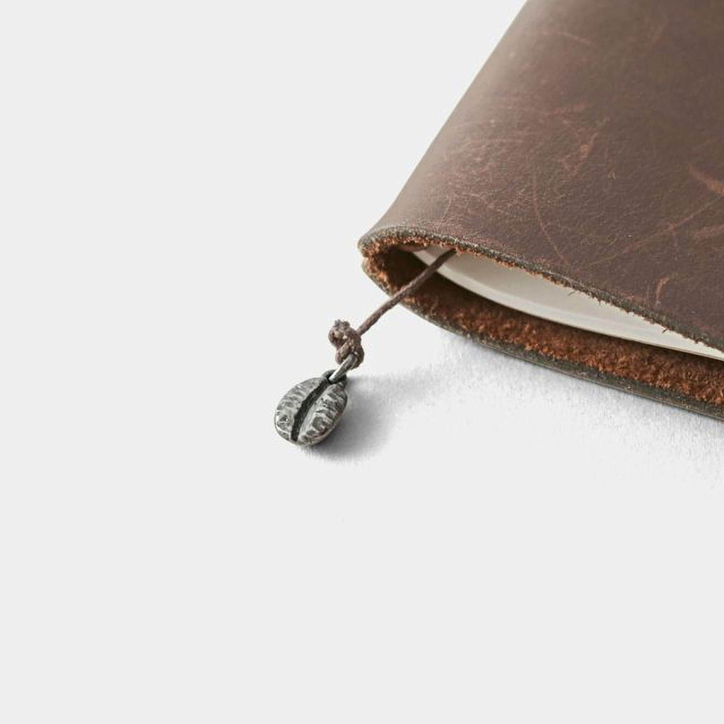 A brown leather-bound notebook with a metal coffee bean charm attached to the page.