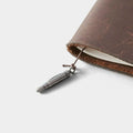 A close-up view of a brown leather-bound notebook with a silver bullet charm attached to the page.