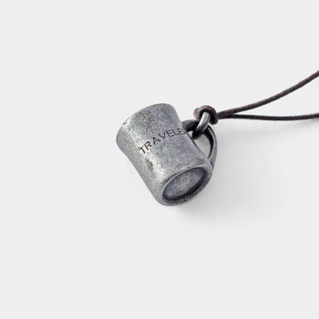 A silver pendant in the shape of a coffee cup with the word "TRAVEL" engraved on it, hanging from a cord.