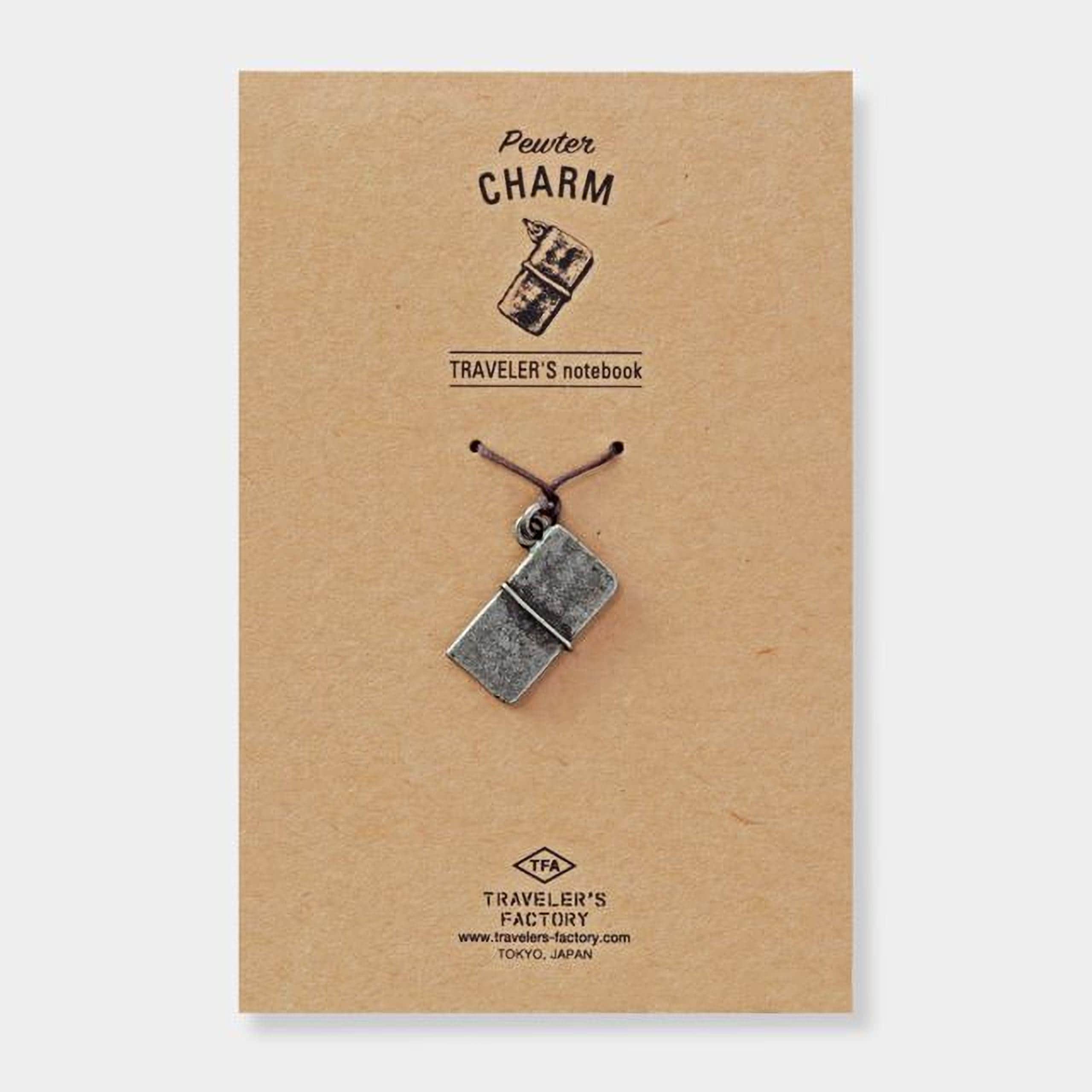 A pewter charm in the shape of a book is attached to a brown paper background, with the text "Pewter Charm" and "TRAVELER'S notebook" printed on it.