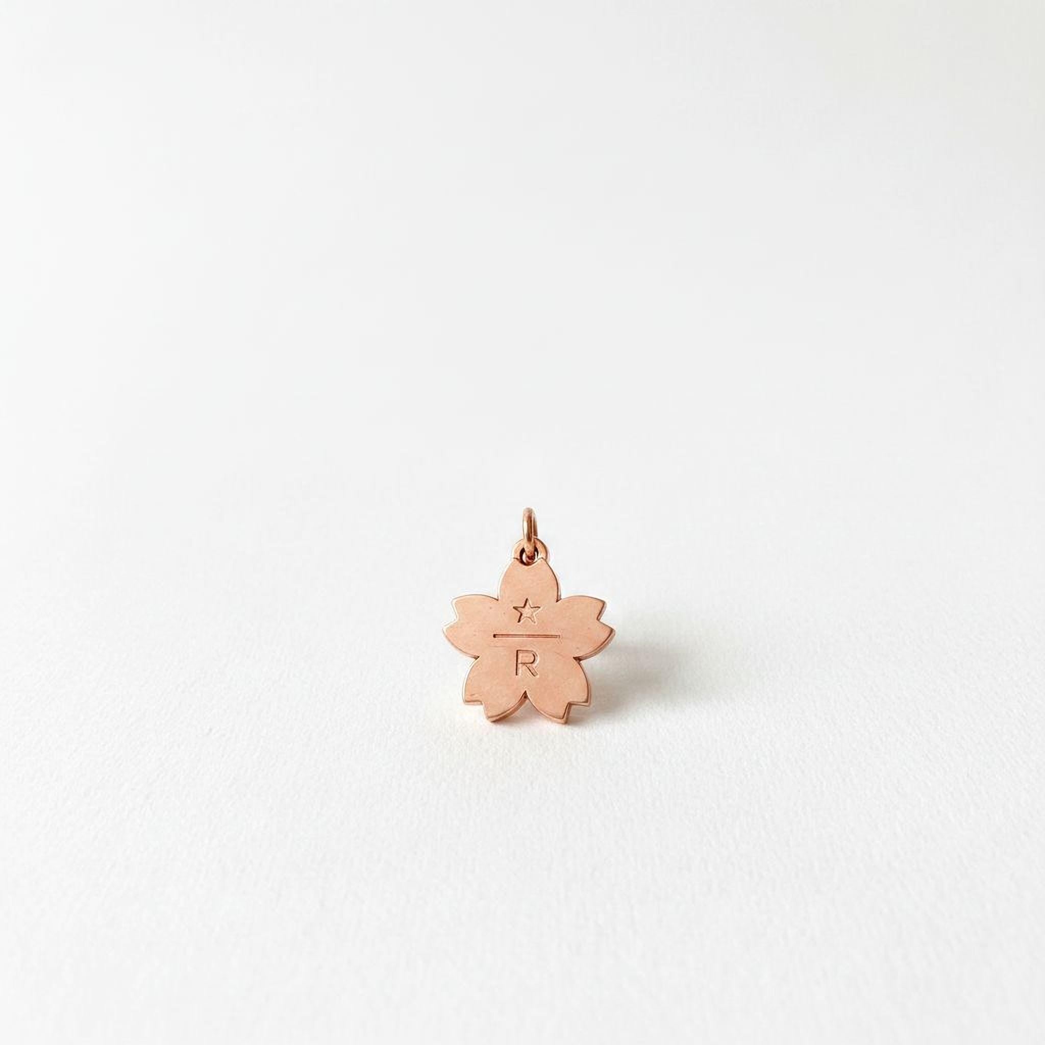 A single, small, metallic charm in the shape of a flower with a letter "R" engraved on it, set against a plain white background.