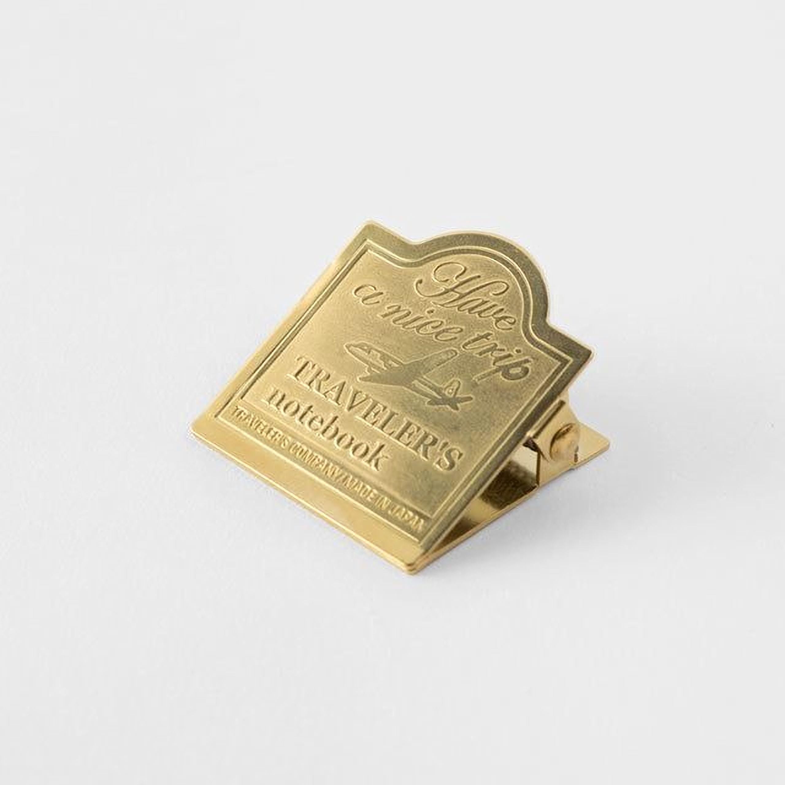 A gold-colored metal clip with embossed text and an image of a plane on it.