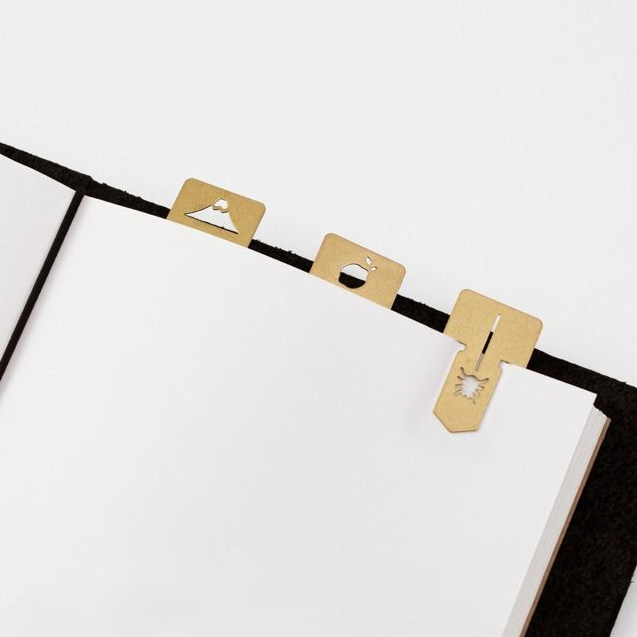 three gold-colored metal bookmarks with various designs, including a mountain, a heart, and a bird, attached to the edge of a white page.