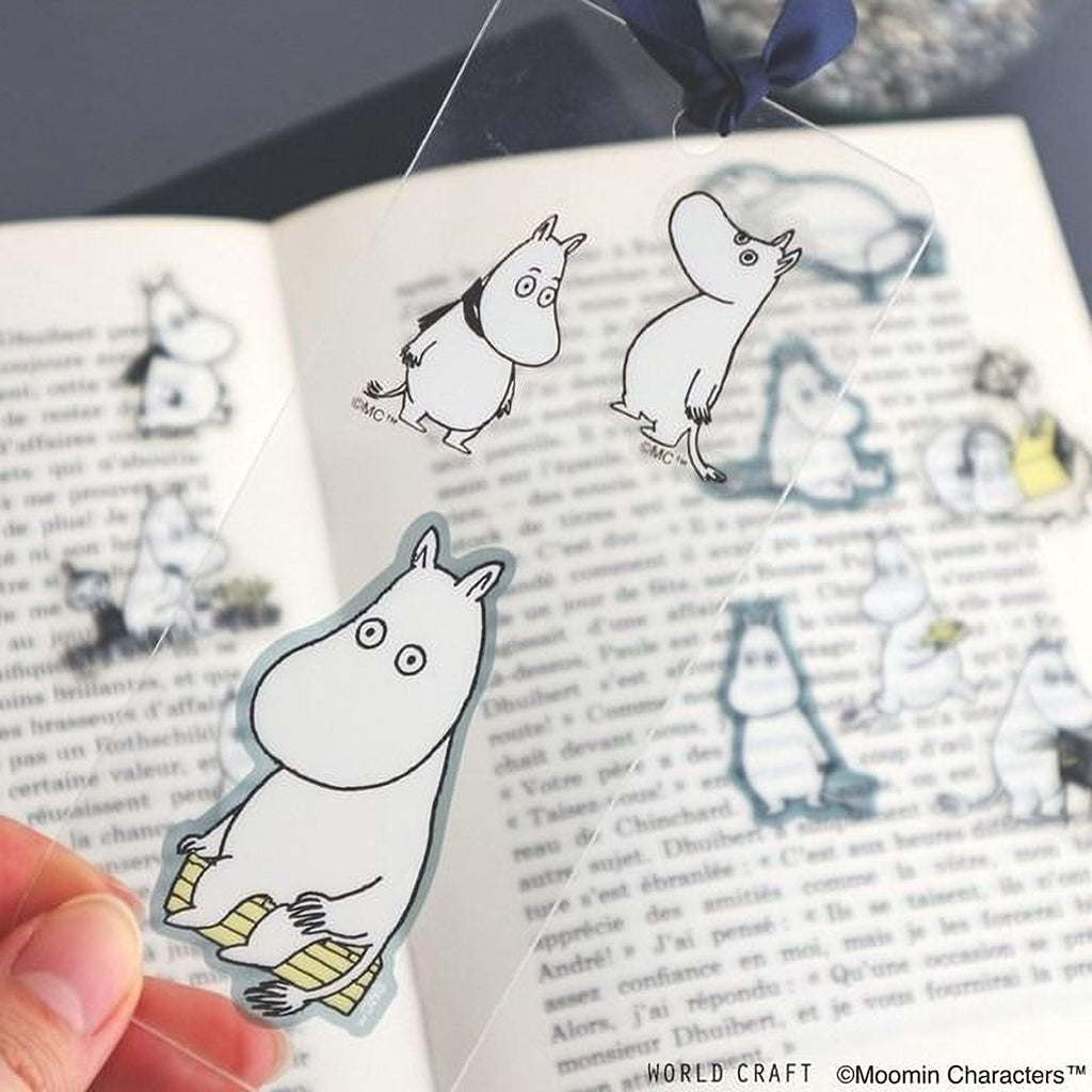 a hand holding a transparent bookmark with illustrations of Moomin characters on it, with a blurred background of text from a book.