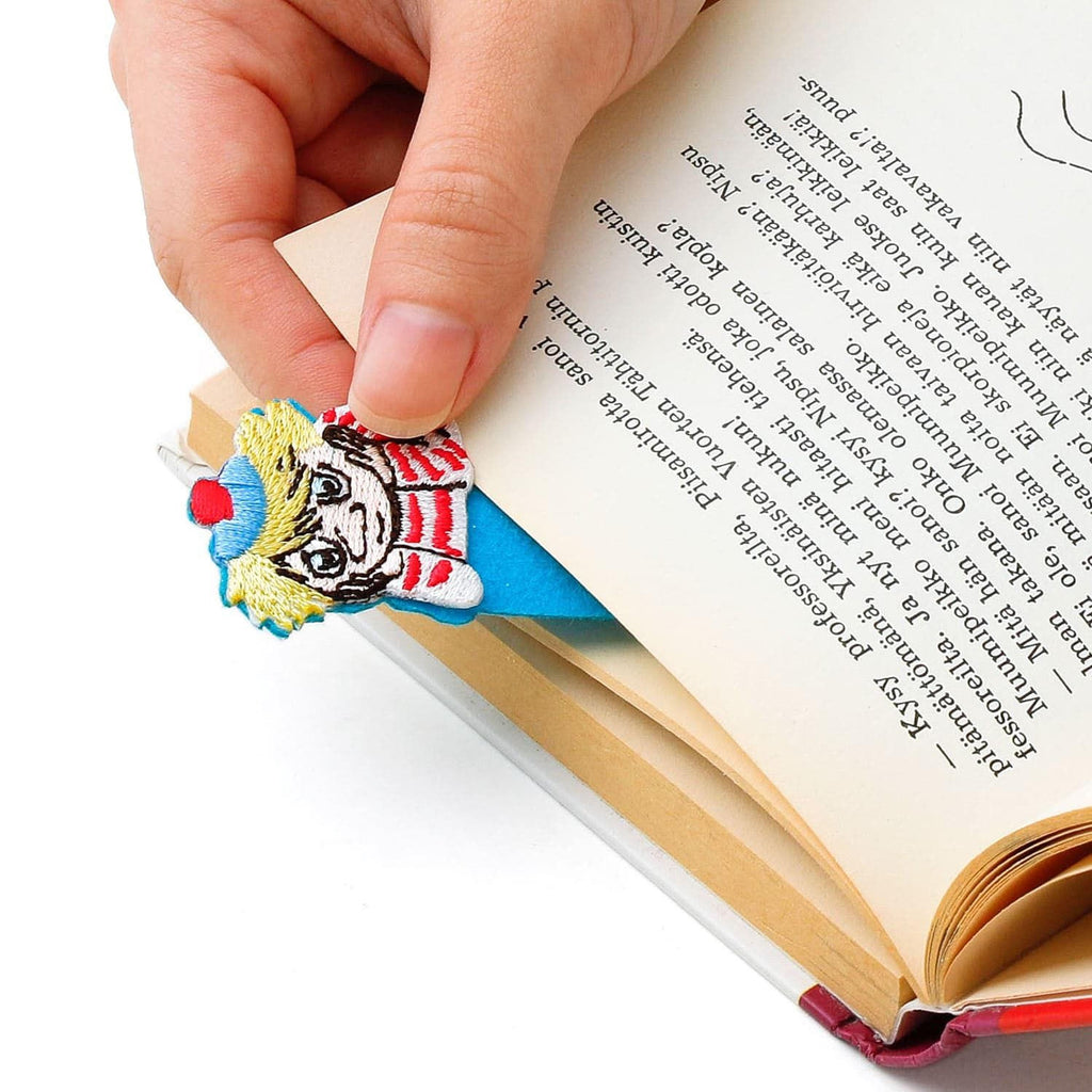 A hand is holding a colorful bookmark shaped like a character, with the bookmark peeking out from between the pages of an open book.