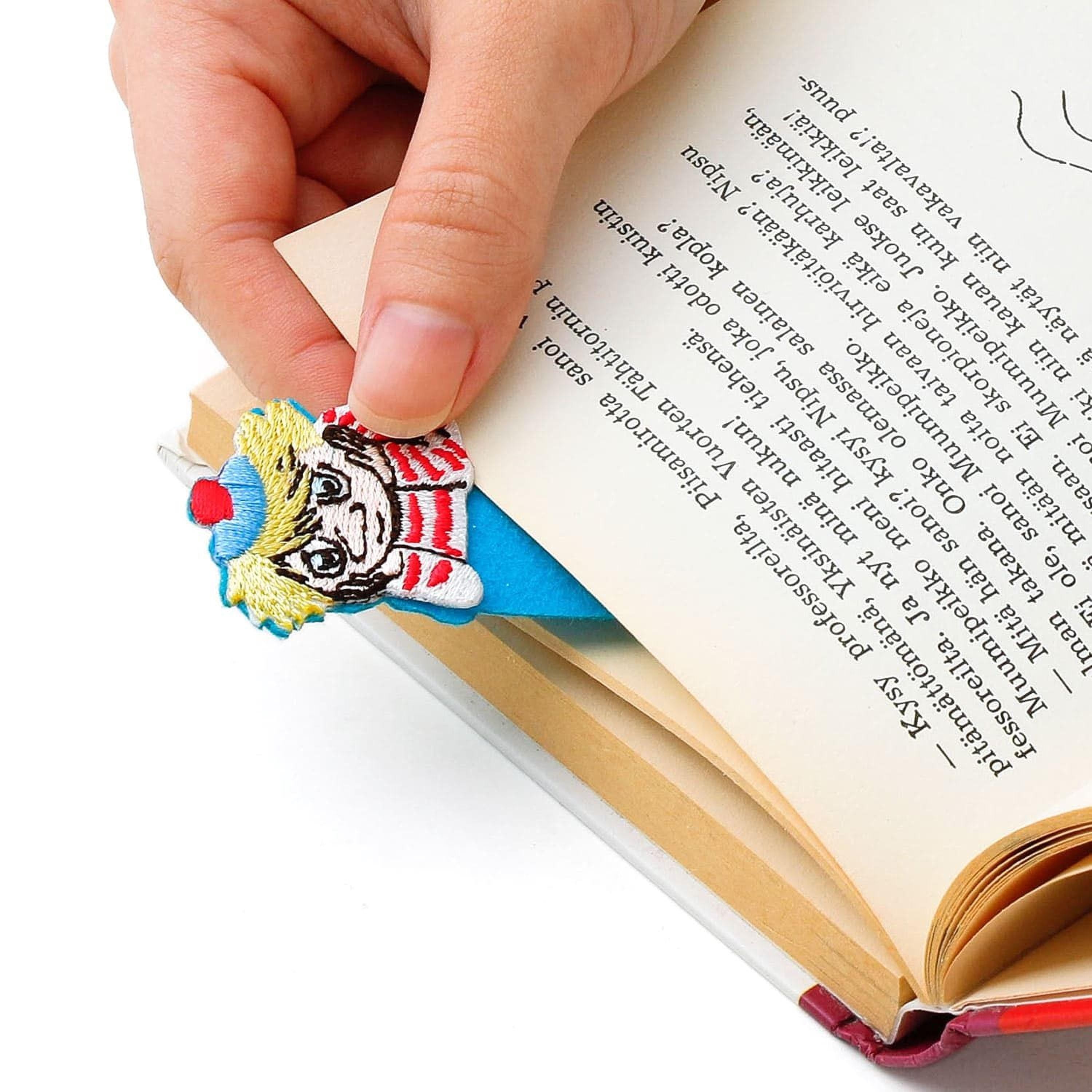 A hand is holding a colorful bookmark shaped like a character, with the bookmark peeking out from between the pages of an open book.