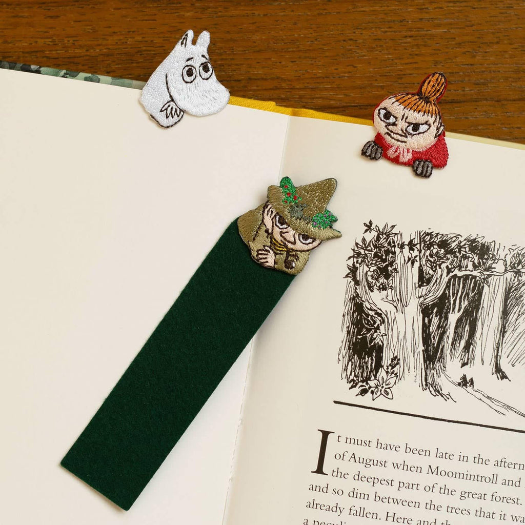 a green bookmark with a whimsical character design, placed on top of an open book with a page illustration of a forest scene.
