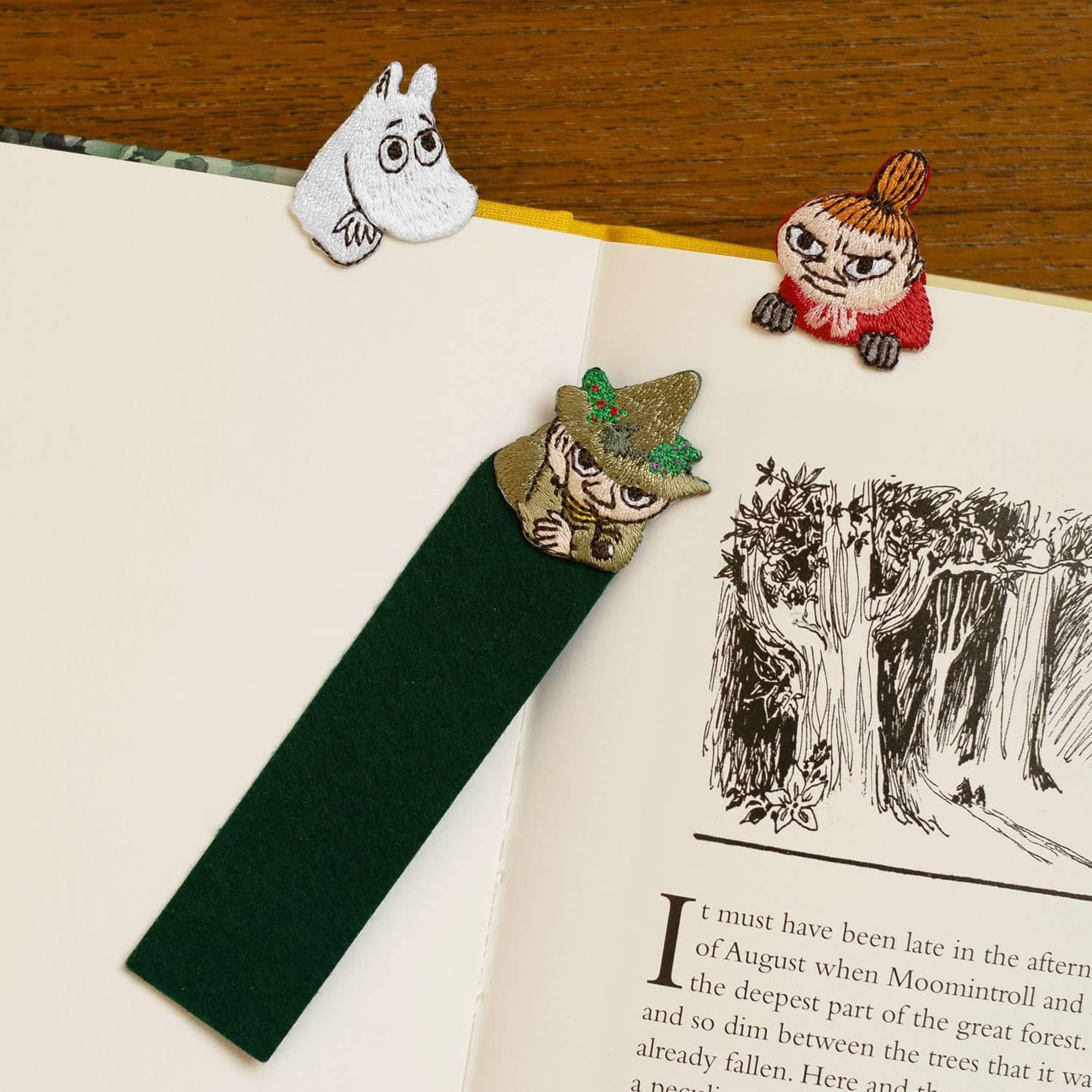 a green bookmark with a whimsical character design, placed on top of an open book with a page illustration of a forest scene.
