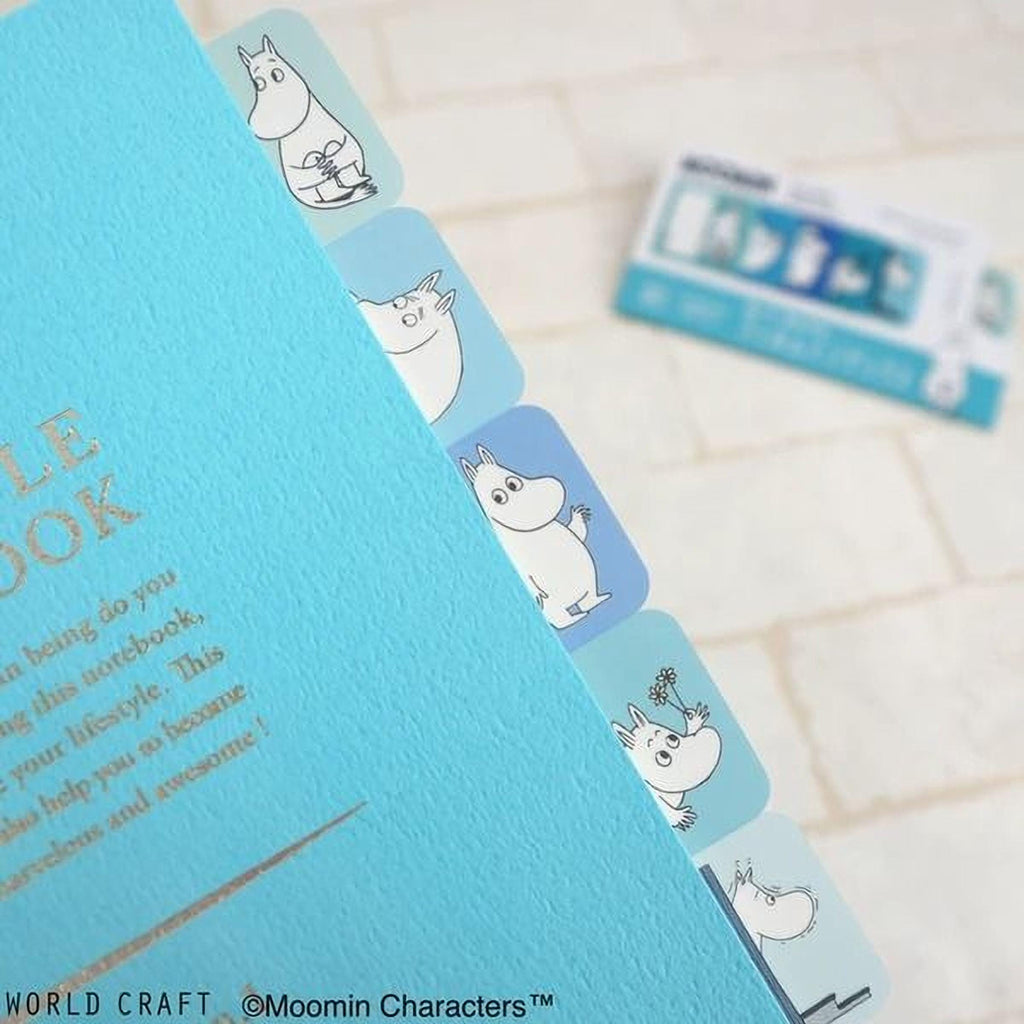 a blue notebook with a playful design of cartoon-style characters on the cover, and a blurred background that includes a card or piece of paper with text and illustrations.