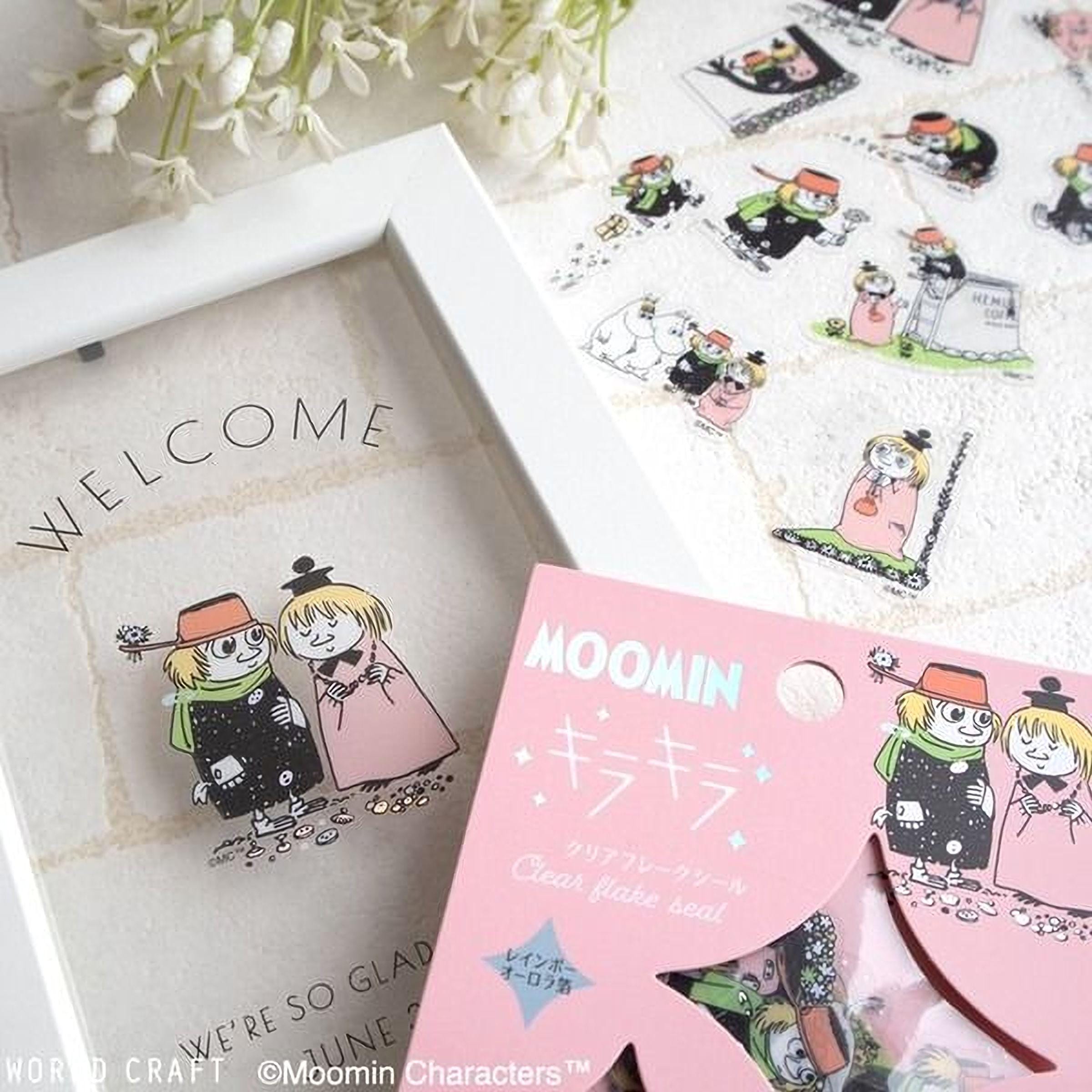 a framed greeting with a "Welcome" message, along with a pink box of Moomin characters stickers. The stickers are illustrated with various cartoon characters, including a girl and a boy. The overall scene appears to be a decorative arrangement with a focus on the greeting and the stickers.