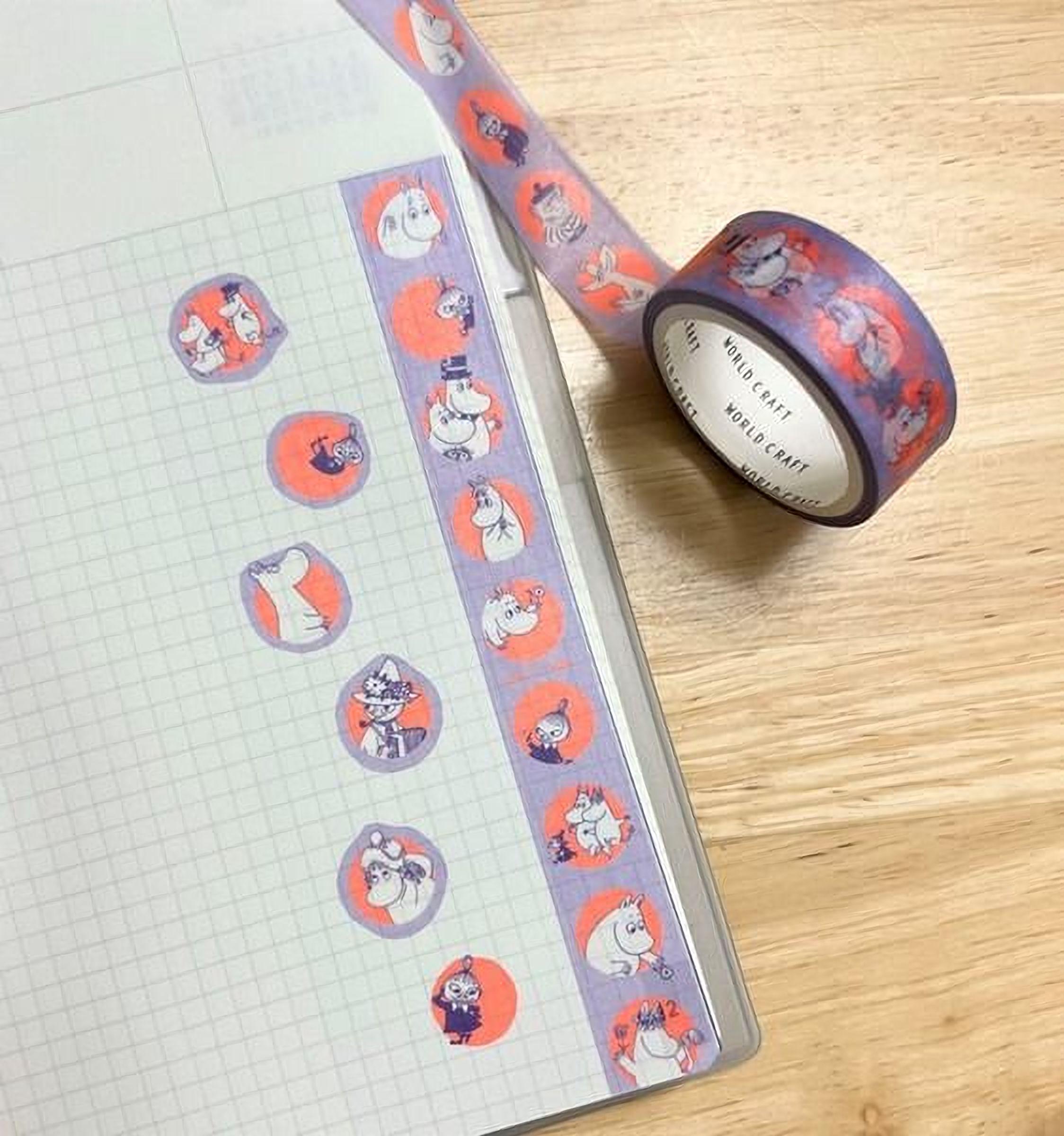 a collection of circular stickers with various cartoon-like illustrations, including a character wearing a top hat and a bowler hat, as well as other whimsical designs. The stickers are arranged on a notebook page, which has a grid pattern. A roll of tape is also visible in the scene, placed next to the notebook.