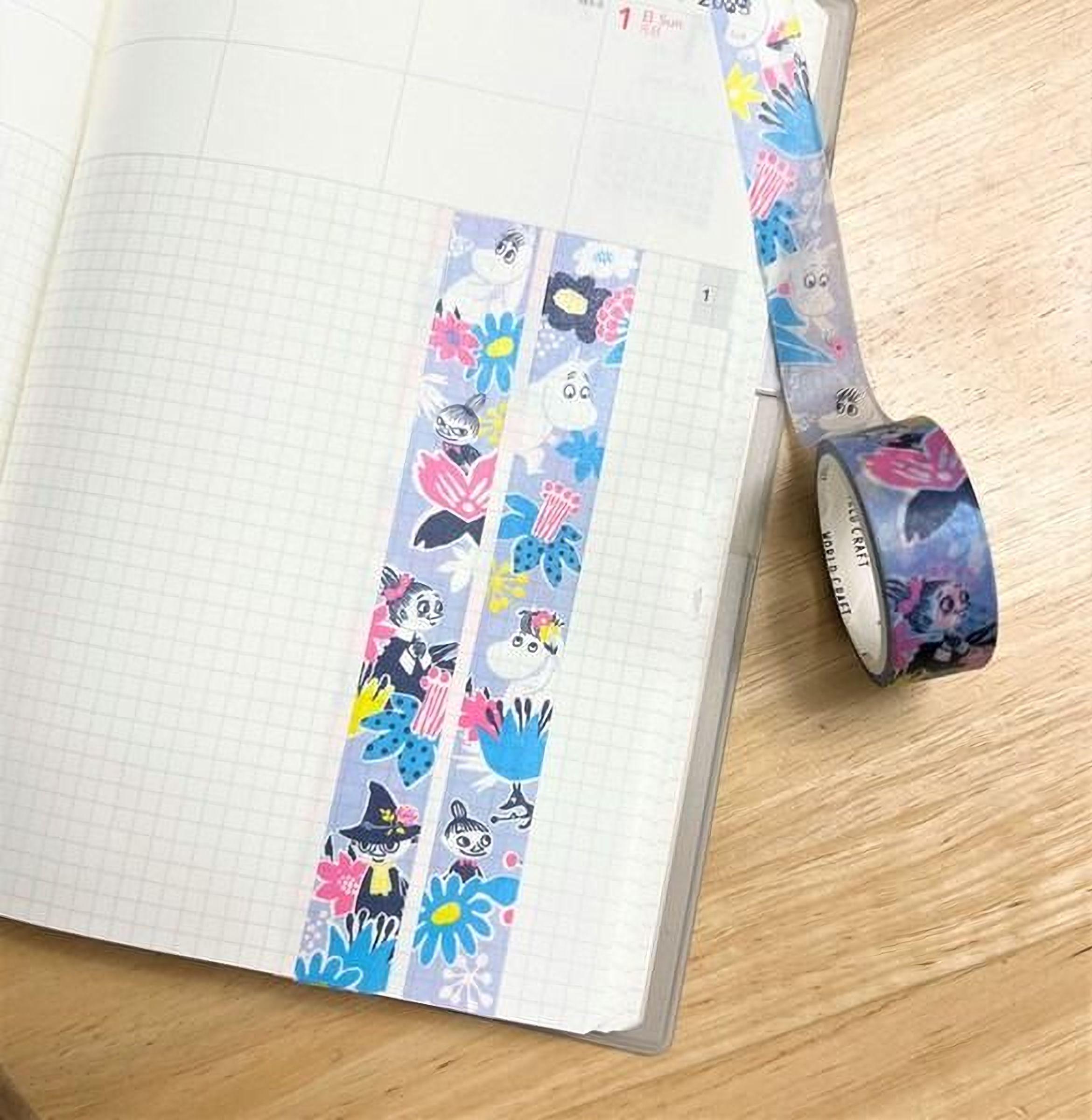 a notebook with a grid pattern and a roll of colorful washi tape with a pattern of cartoon characters on it, placed on a wooden surface.