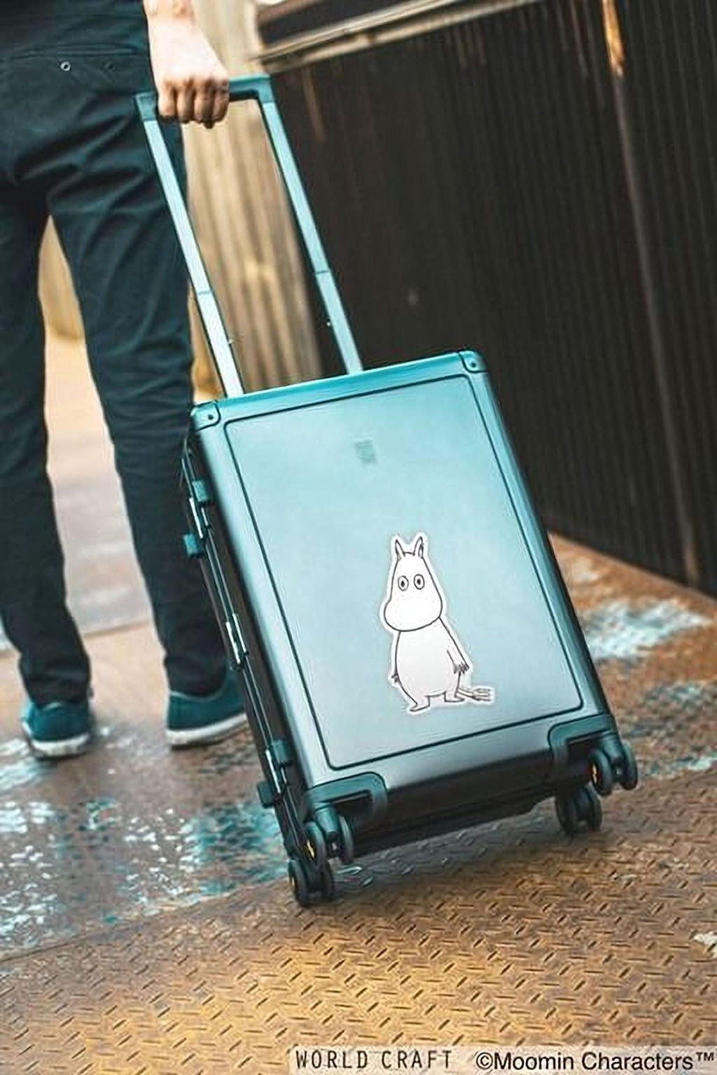 A person is pulling a suitcase with a sticker of a character on it, standing on a metal surface.