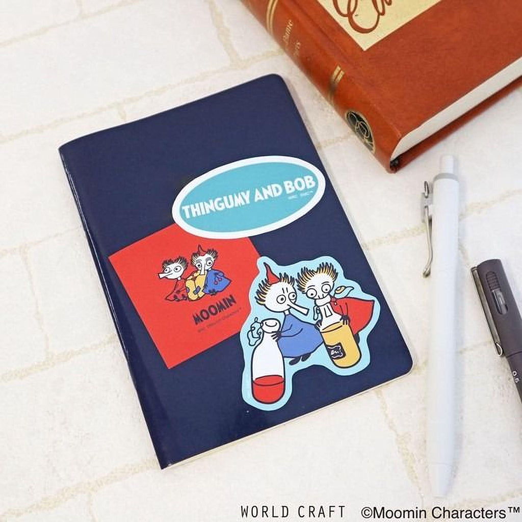 a blue notebook with a cartoon character sticker, a red book, and a pen on a white surface.