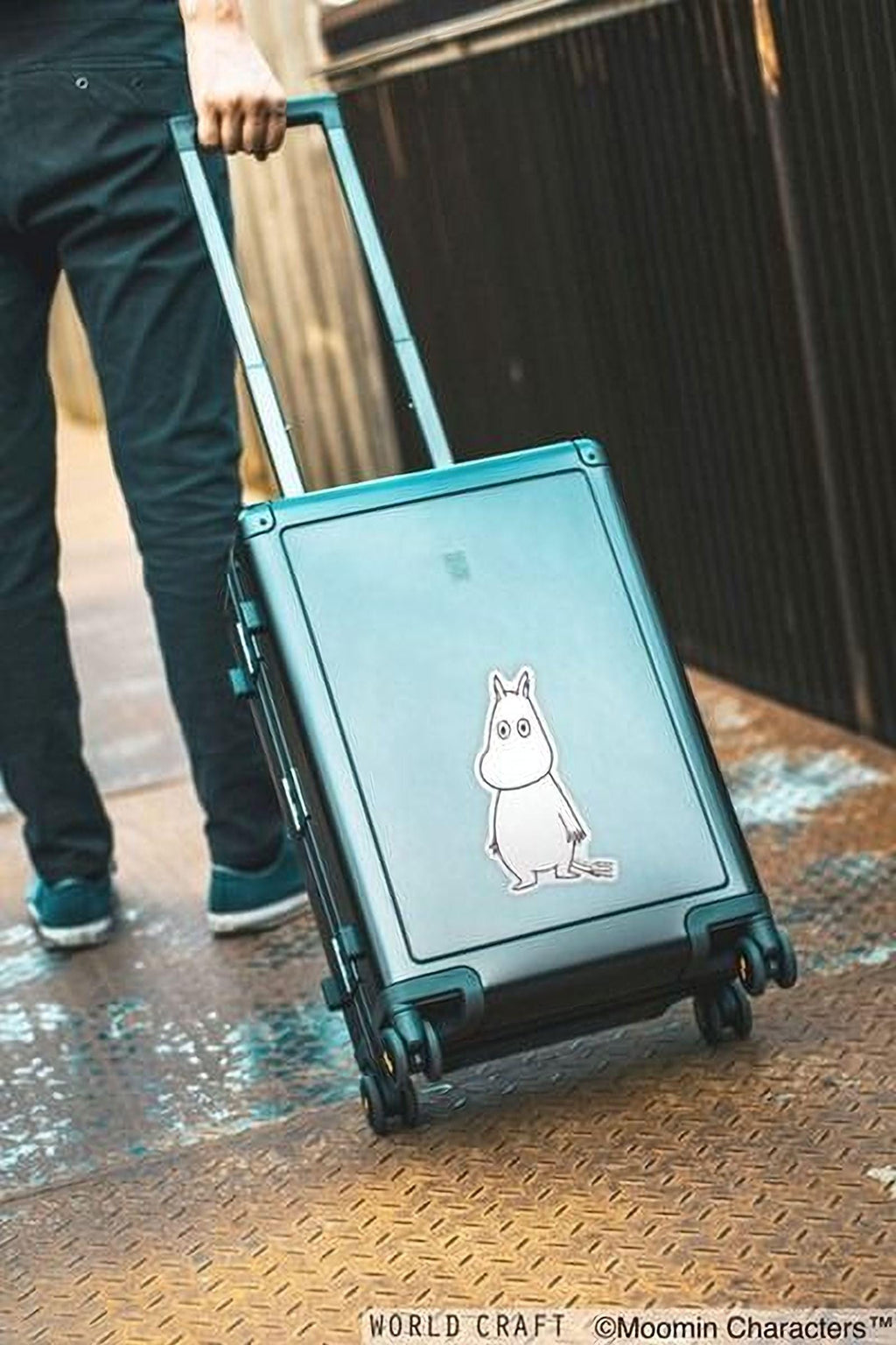 A person is pulling a suitcase with a sticker of a character on it, standing on a metal surface.