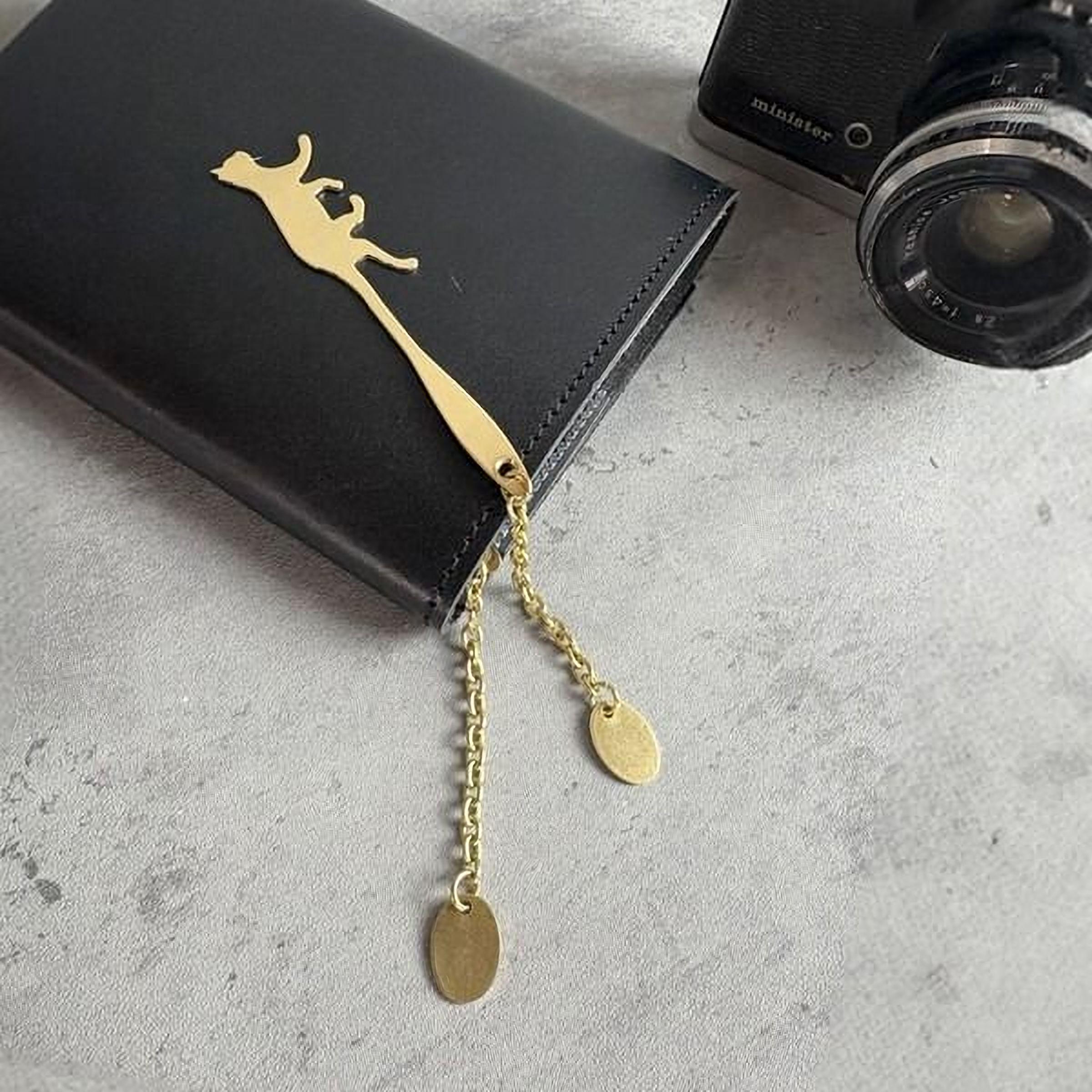 a black wallet with a gold cat-shaped bookmark attached to it, placed on a gray surface next to a camera.
