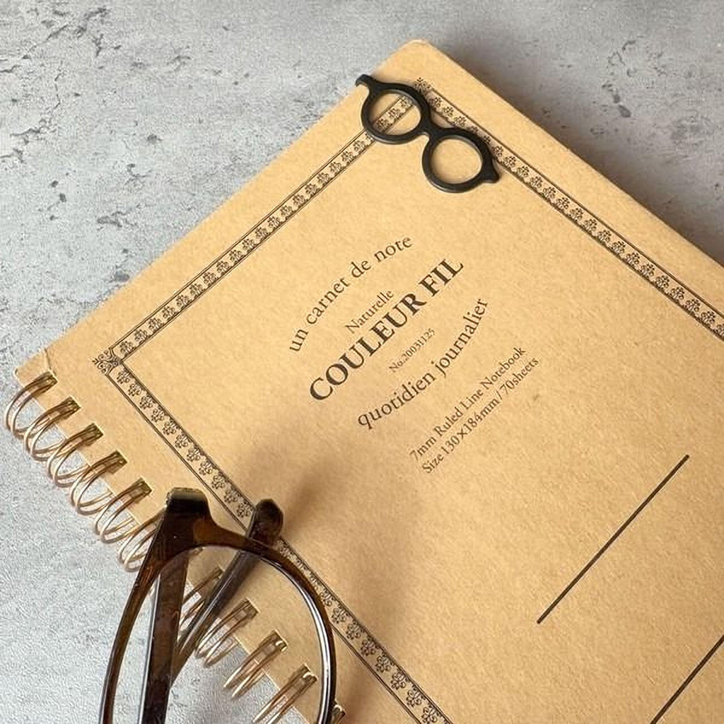 A spiral-bound notebook with a pair of glasses resting on top of it, placed on a gray surface.
