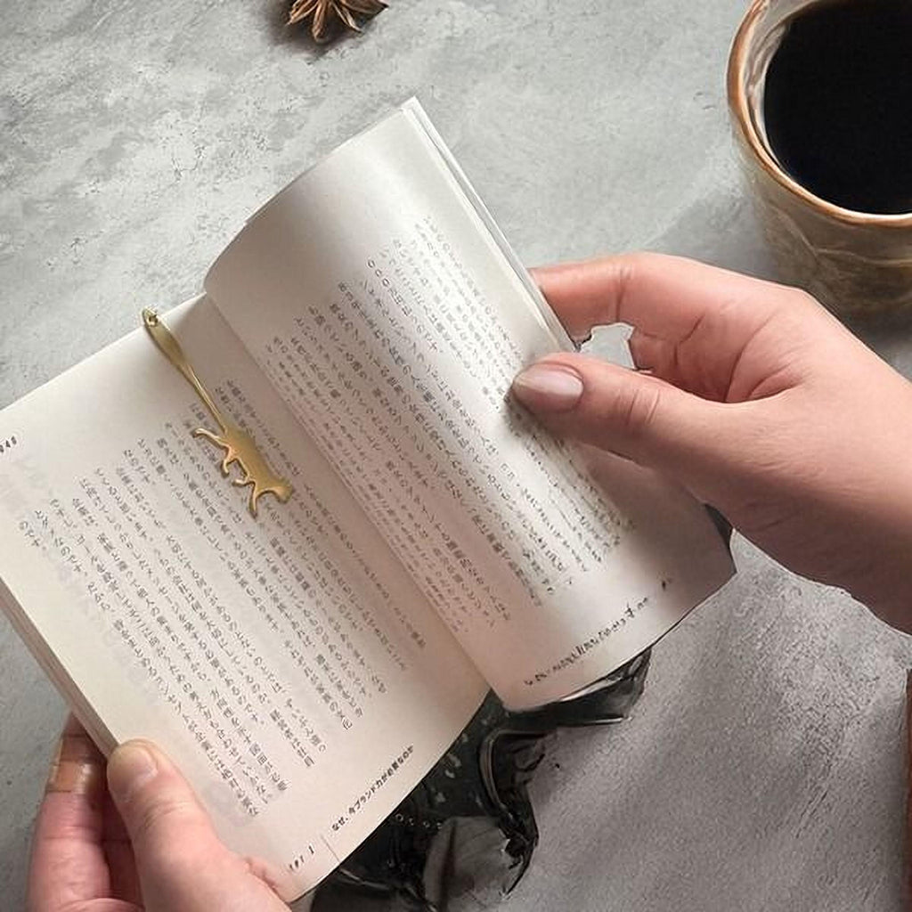 A person's hands holding an open book with a gold bookmark shaped like a cat.