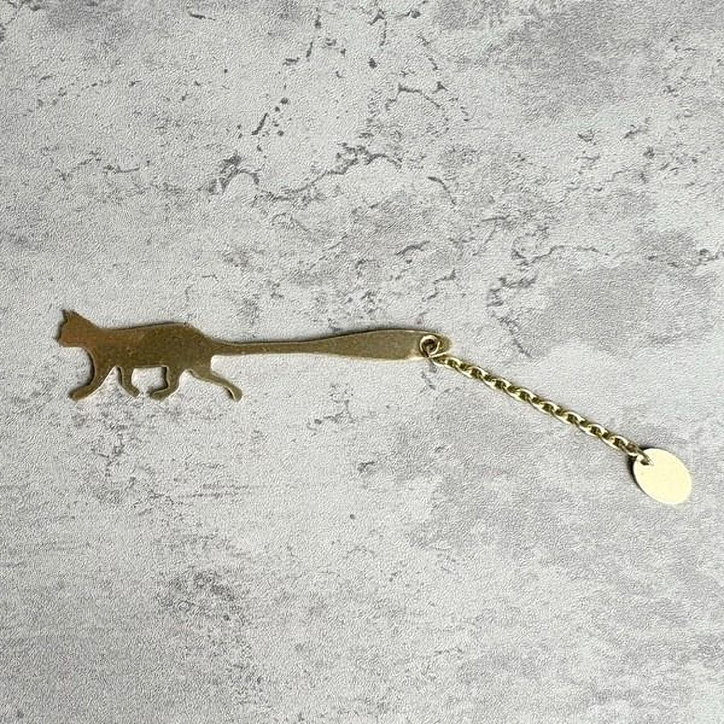 A gold-colored metal keychain in the shape of a cat is shown on a textured gray surface.