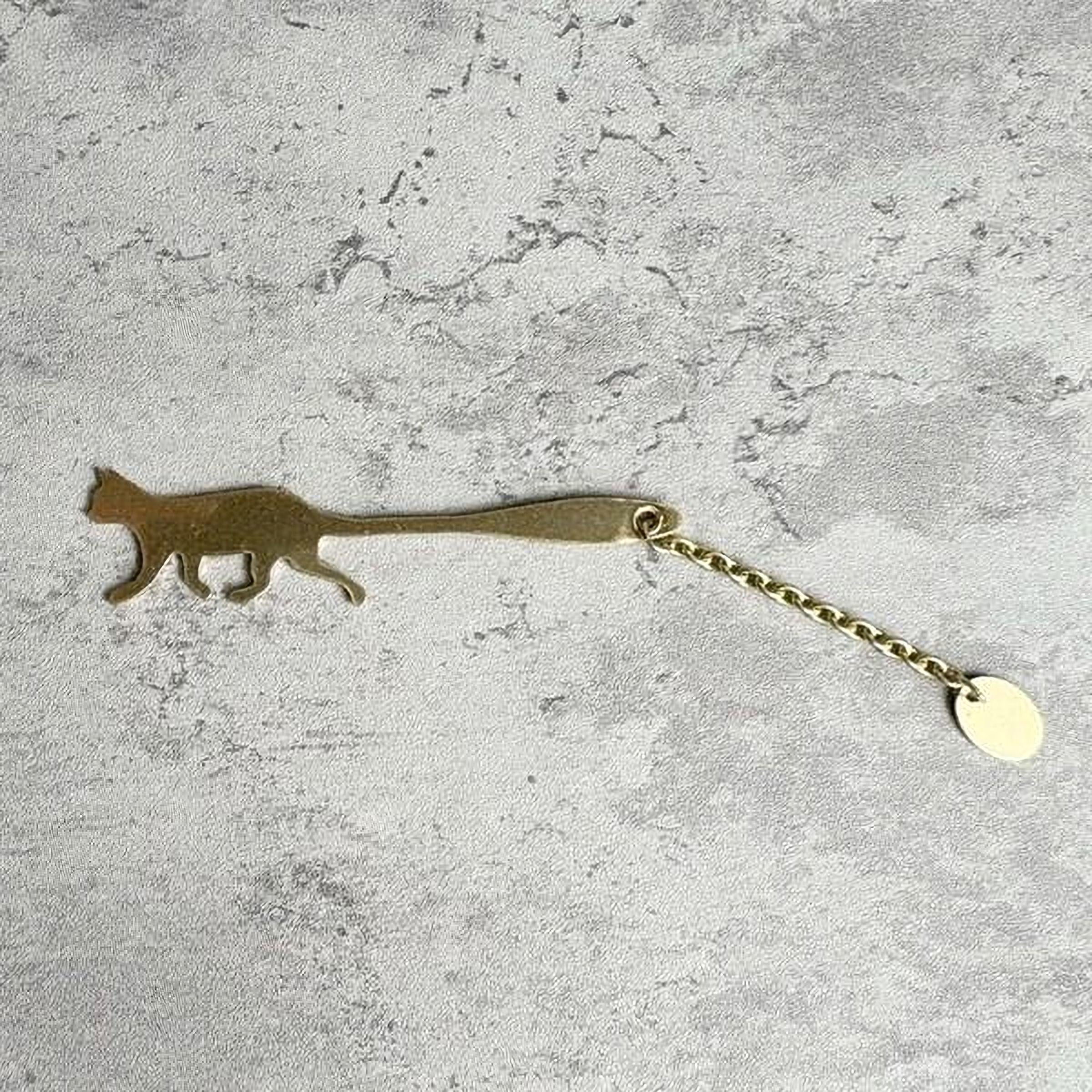 A gold-colored metal keychain in the shape of a cat is shown on a textured gray surface.