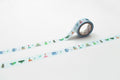 A roll of blue and white patterned tape with various objects such as cups, bowls, and plants printed on it, lying on a white surface.