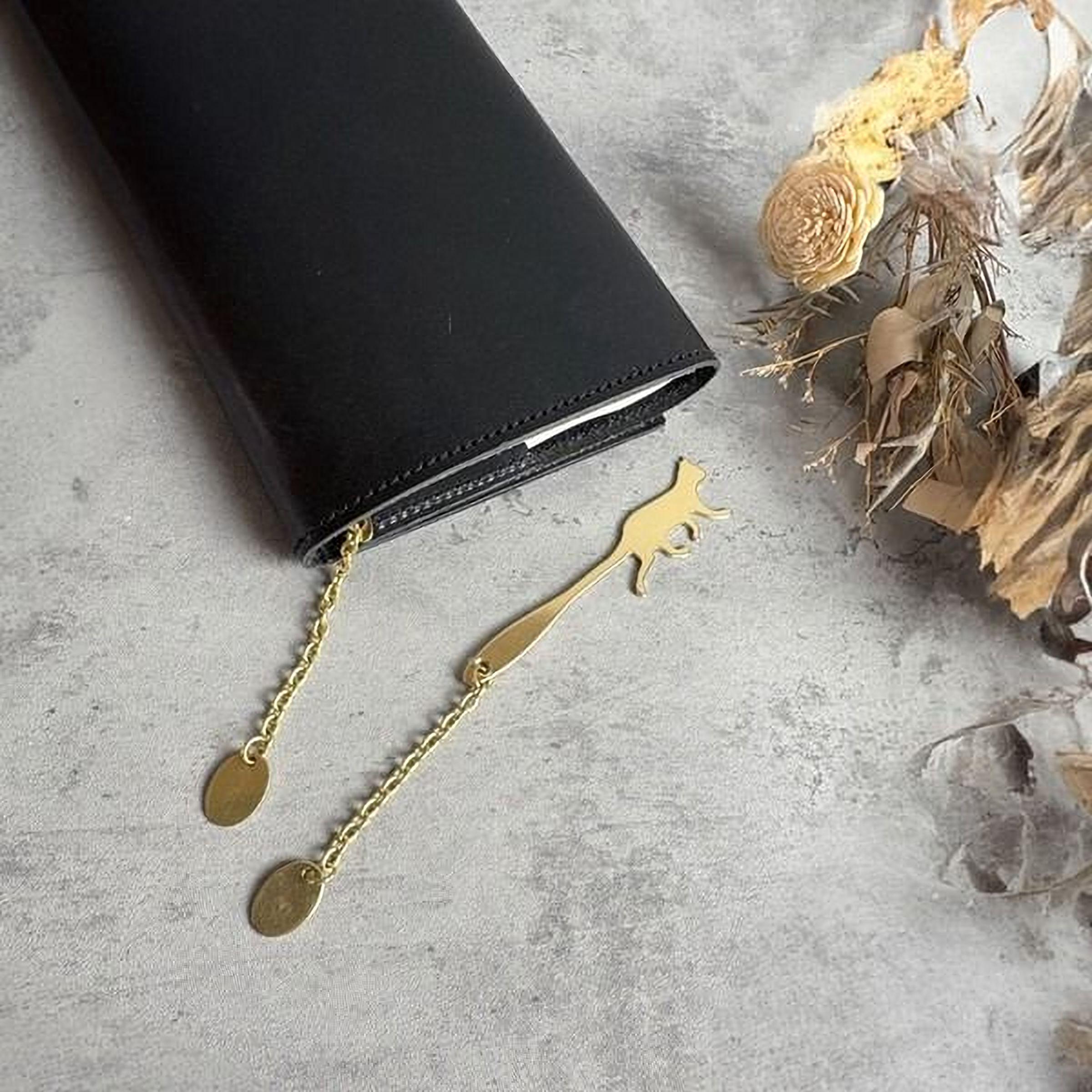 A black wallet or purse with a gold cat-shaped bookmark attached to it, along with some dried flowers and a dried leaf on a gray surface.
