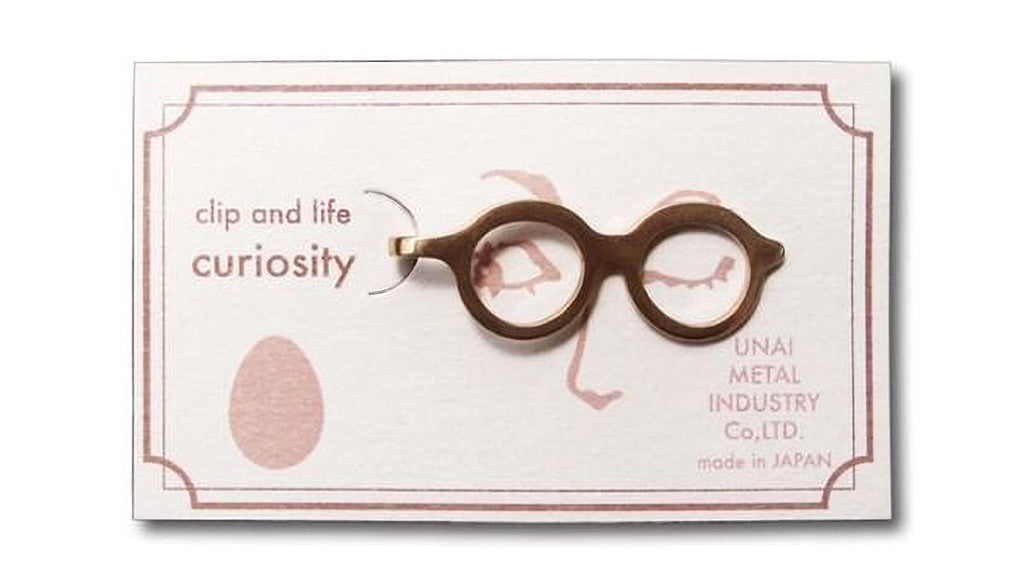 a pair of gold-colored glasses with a face design on the frame, placed on a white background with text and a small pink circle.