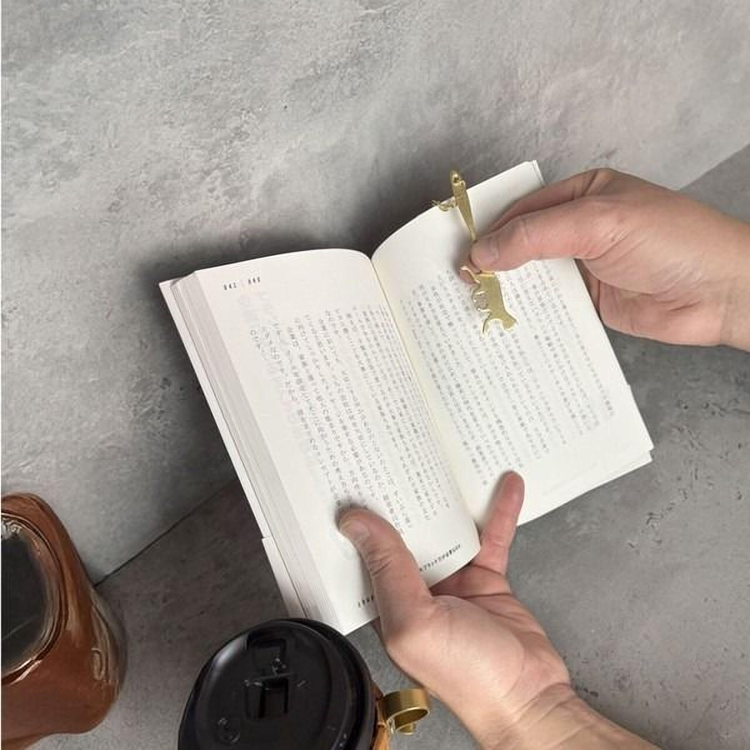 A person's hands holding an open book with a gold bookmark attached to it, resting on a gray surface.
