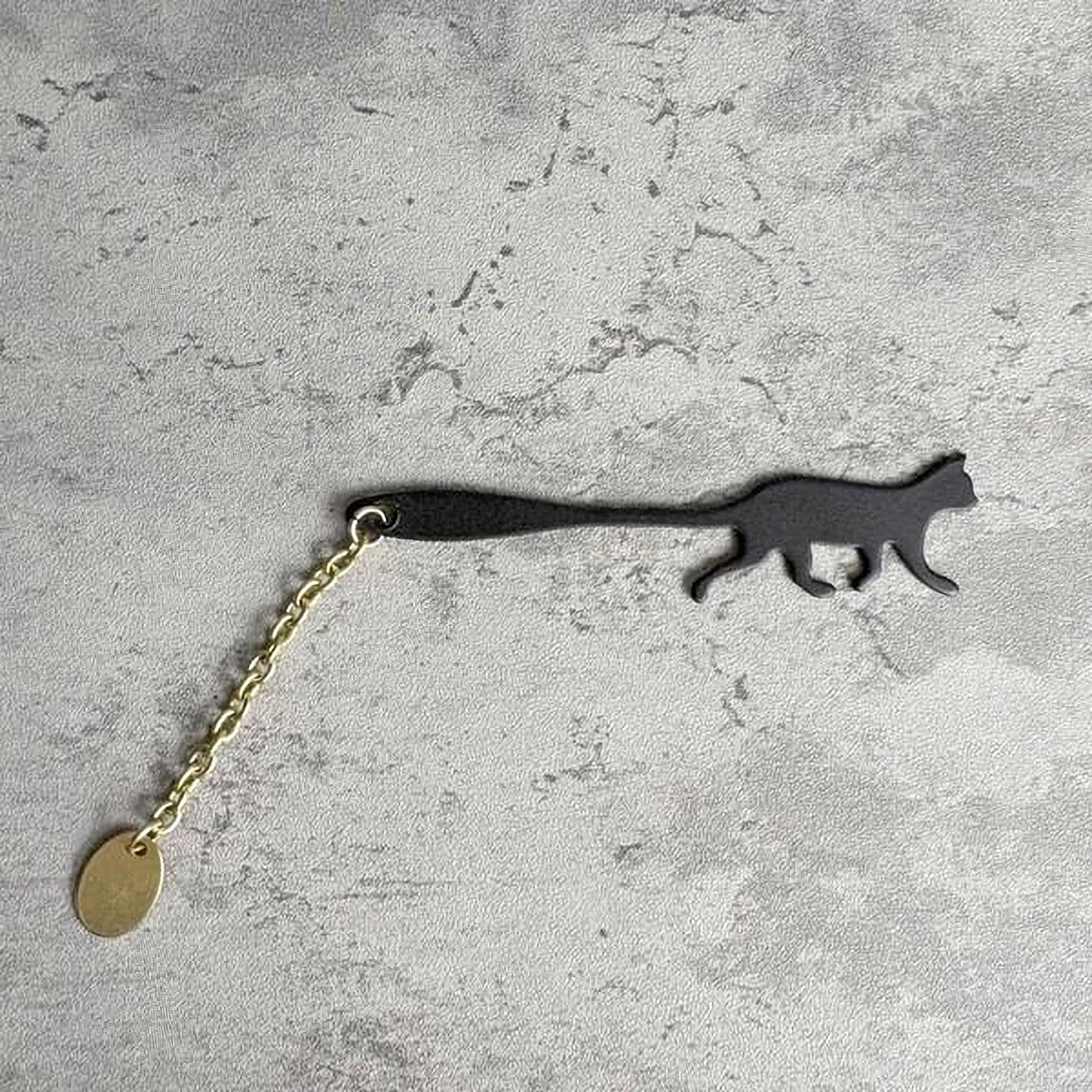 A black cat-shaped keychain with a gold chain attached to it, lying on a textured gray surface.