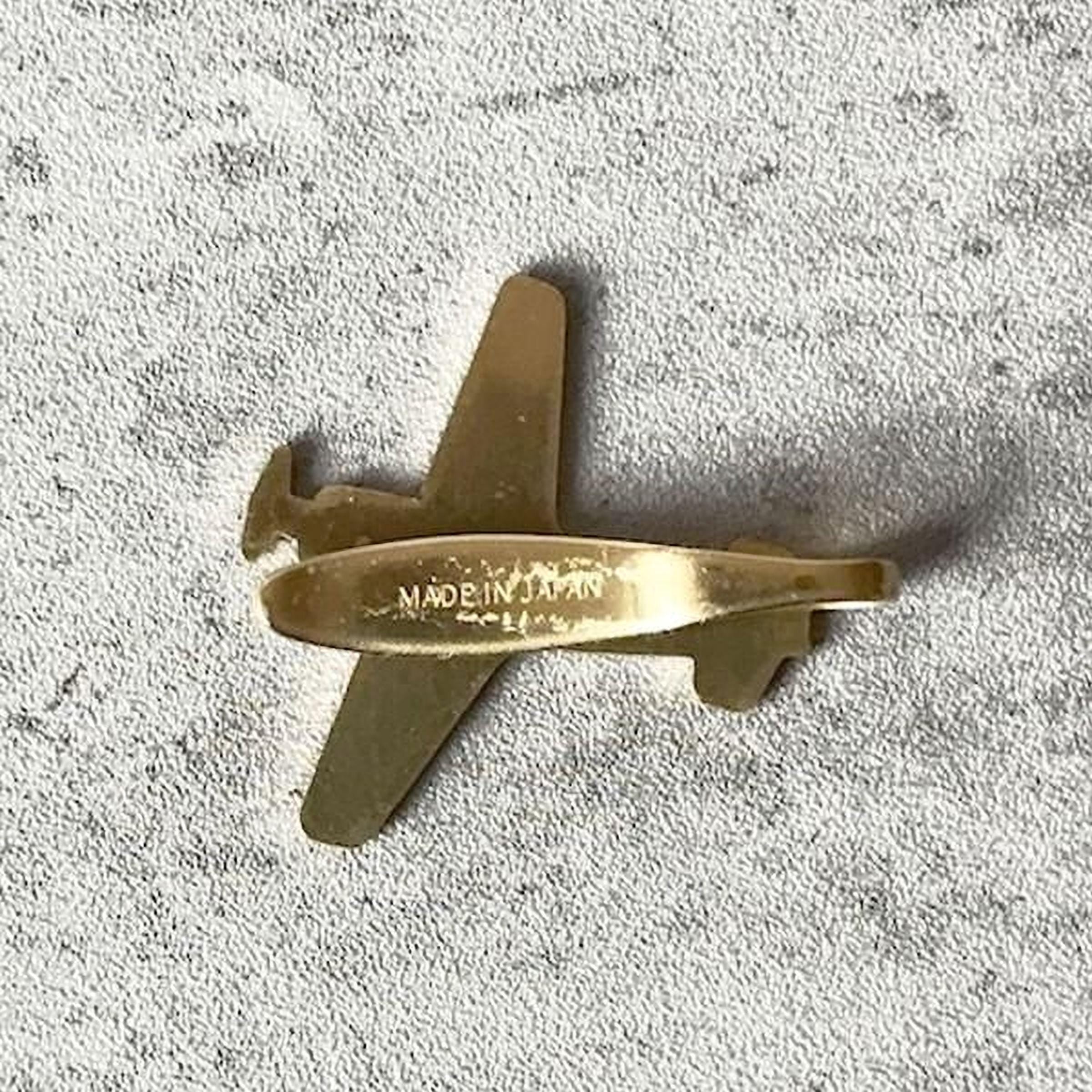 A small gold-colored airplane pin is shown against a textured gray background.