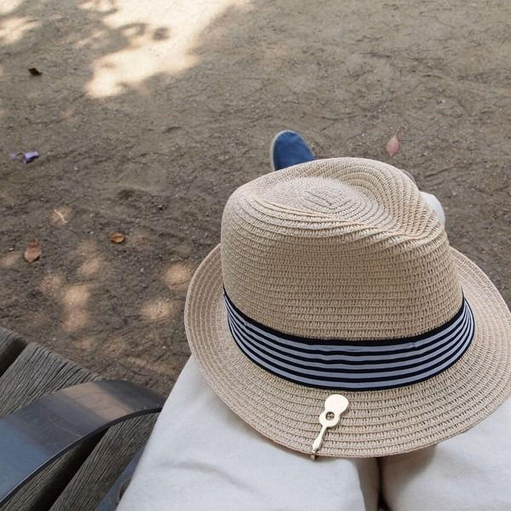 A person wearing a straw hat with a black and white striped band is sitting on a wooden bench in a park, with fallen leaves on the ground around them.