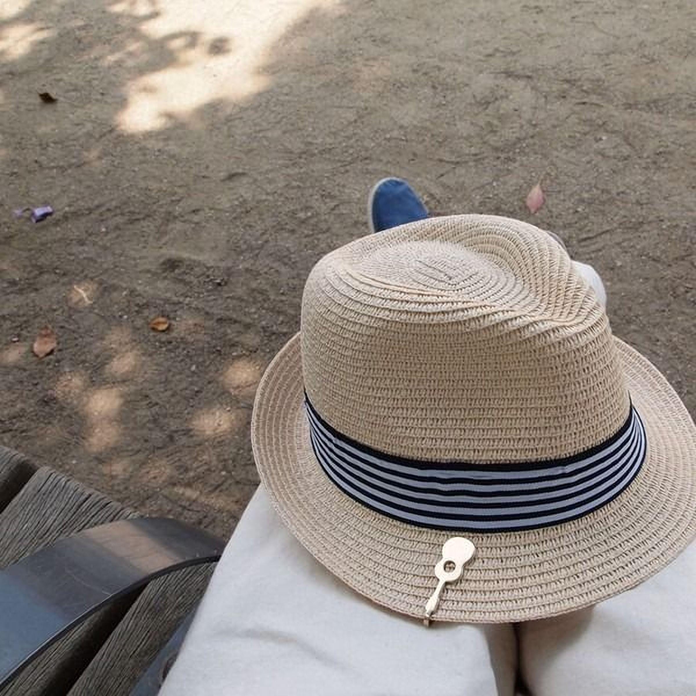 A person wearing a straw hat with a black and white striped band is sitting on a wooden bench in a park, with fallen leaves on the ground around them.