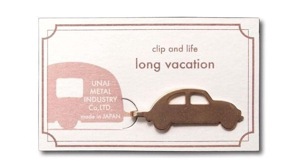 a small, bronze-colored car keychain shaped like a Volkswagen Beetle, with the text "clip and life long vacation" and "UNAF Metal Industry Co., Ltd. made in JAPAN" printed on it.