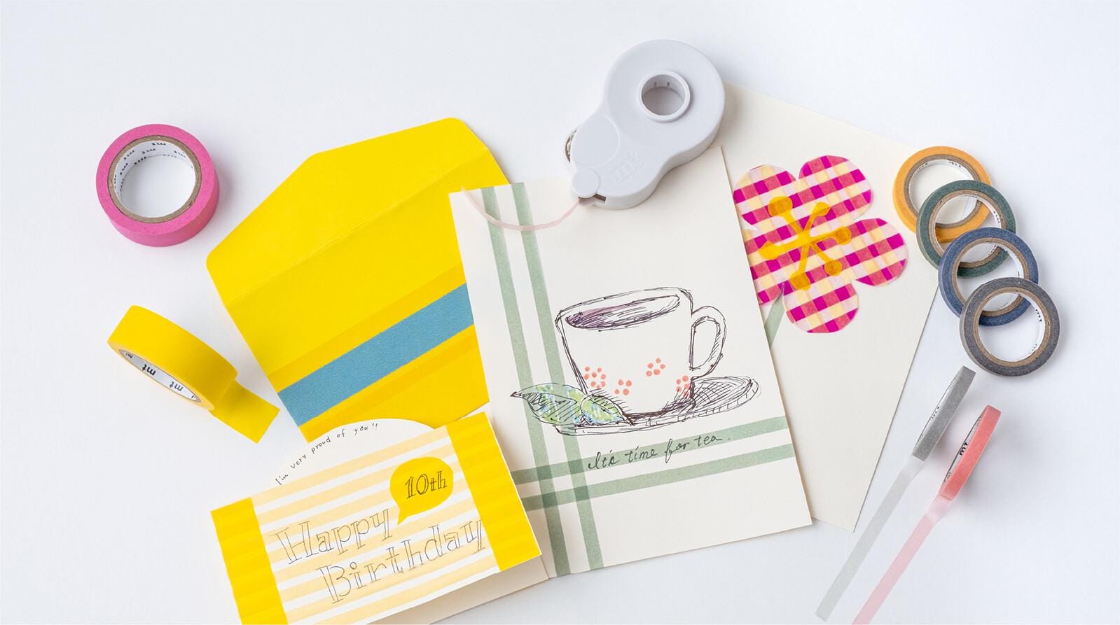 various craft supplies, including a yellow envelope, a white envelope, a cup, and several rolls of tape and markers, all arranged on a white background.