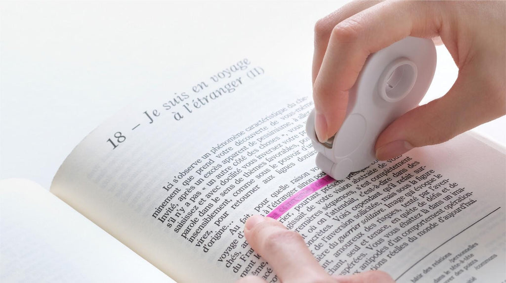 A person's hand is holding a white object, possibly a stapler or a highlighter, and is marking or highlighting text on an open book.